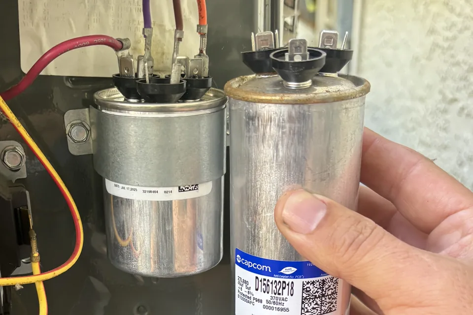 Alpha Mechanical technician holding a failed dual run capacitor with a swollen, domed and corroded top beside its flat-topped replacement wired into the condenser