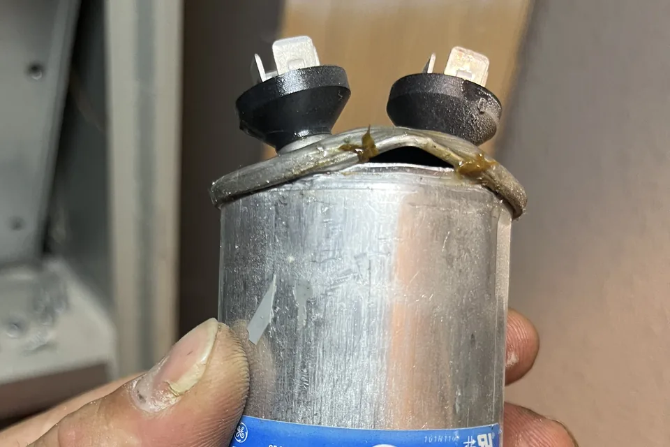 Failed GE run capacitor pulled from a Sacramento air conditioner, its top seam split open with amber dielectric leaking out between the two terminals