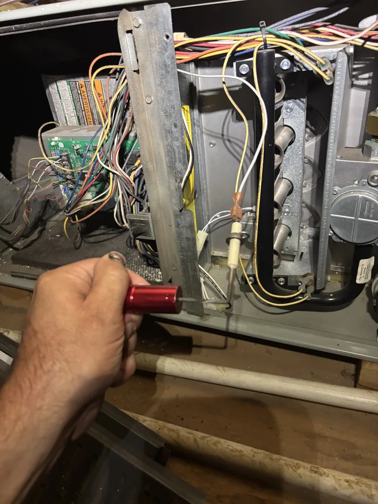 Cleaning the flame sensor rod on a gas furnace burner assembly during an Alpha Mechanical service call: the rod sits beside the hot surface igniter, with the row of inshot burners, the gas valve and the control board wiring in view