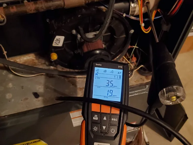 Testo 310 combustion analyzer with its probe in a furnace test port on an Alpha Mechanical diagnostic, the display showing a carbon monoxide reading in parts per million beside the inducer motor