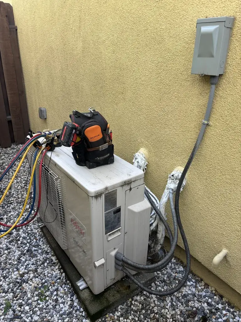 The outdoor unit of a mini-split heat pump beside a stucco wall, with gauge hoses connected and a tool bag on top