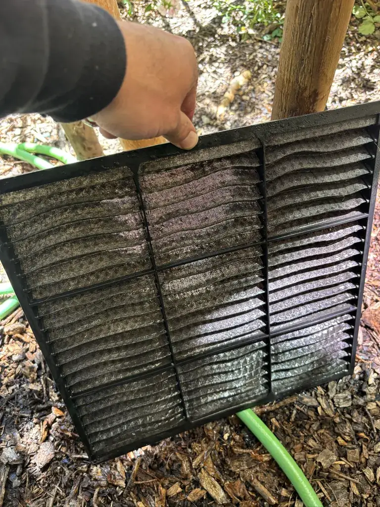 A washable filter from a recessed ceiling unit of a mini-split heat pump, held up outdoors, its mesh grey with dust