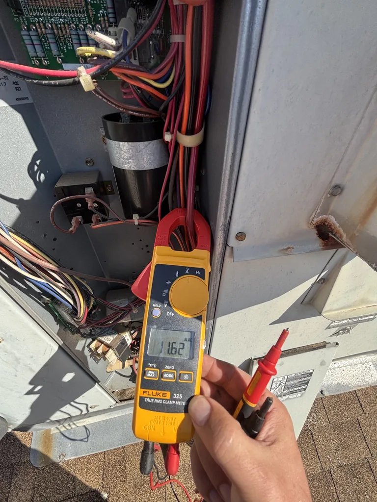 A Fluke 325 clamp meter clamped on a red compressor conductor inside the electrical compartment of a rooftop packaged AC unit, reading 11.62 amps, with the dual-run capacitor, contactor, and control board in view