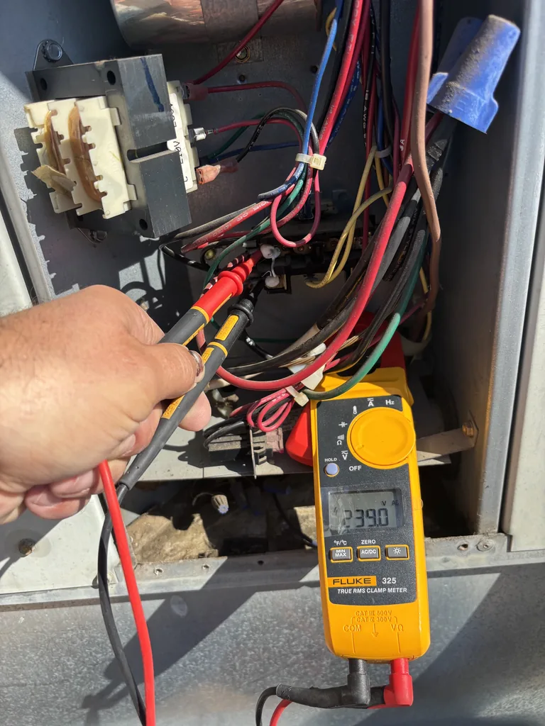 A Fluke 325 clamp meter with test leads on the line terminals of an AC contactor, reading 239.0 volts, with the 24-volt control transformer and wiring harness in the electrical compartment