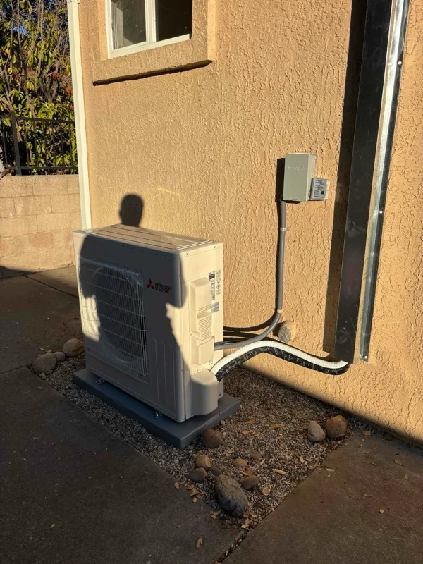 Alpha Mechanical job photo: a Mitsubishi mini split outdoor unit on a composite pad against a stucco wall, with the insulated line set, condensate drain and control cable bundled in a short run through the wall and a weatherproof disconnect mounted above it