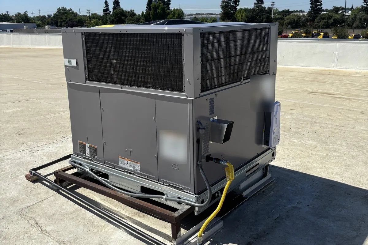 A rooftop package unit set on a steel frame on a Sacramento commercial roof, with its flexible gas connector and electrical conduit, trees and parked cars beyond the parapet