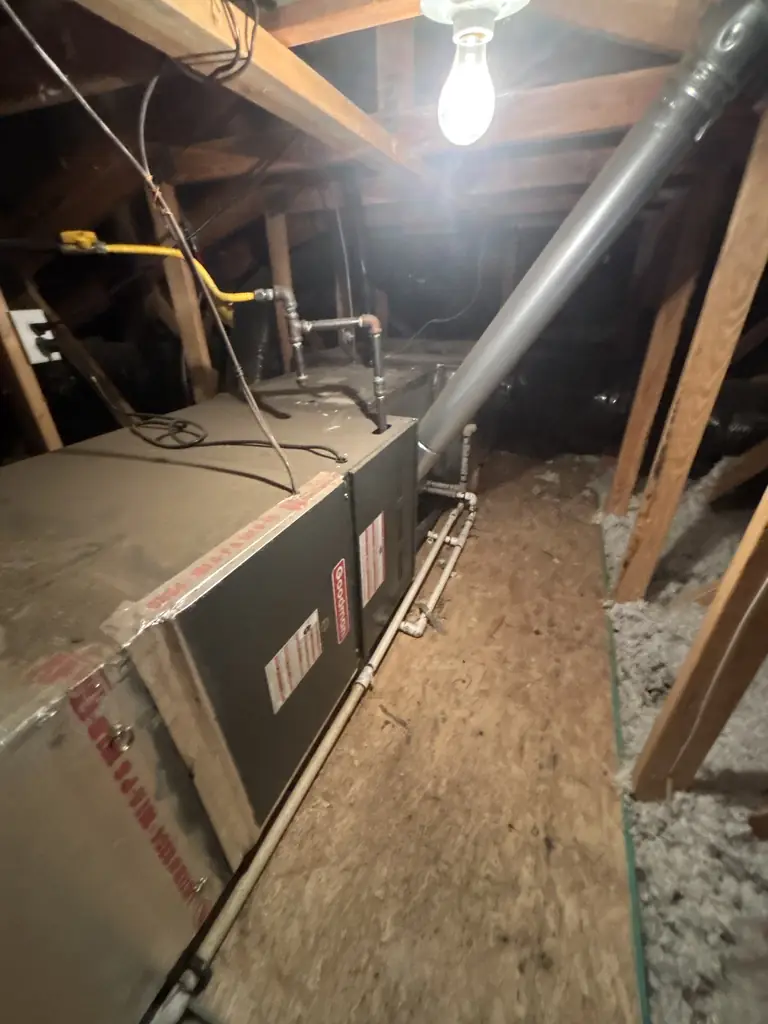 Before: a Goodman gas furnace lying horizontally in a Fair Oaks attic on an Alpha Mechanical service call in June 2026, with a yellow flexible gas connector, a metal flue pipe rising through the roof framing, and white PVC condensate lines along the floor
