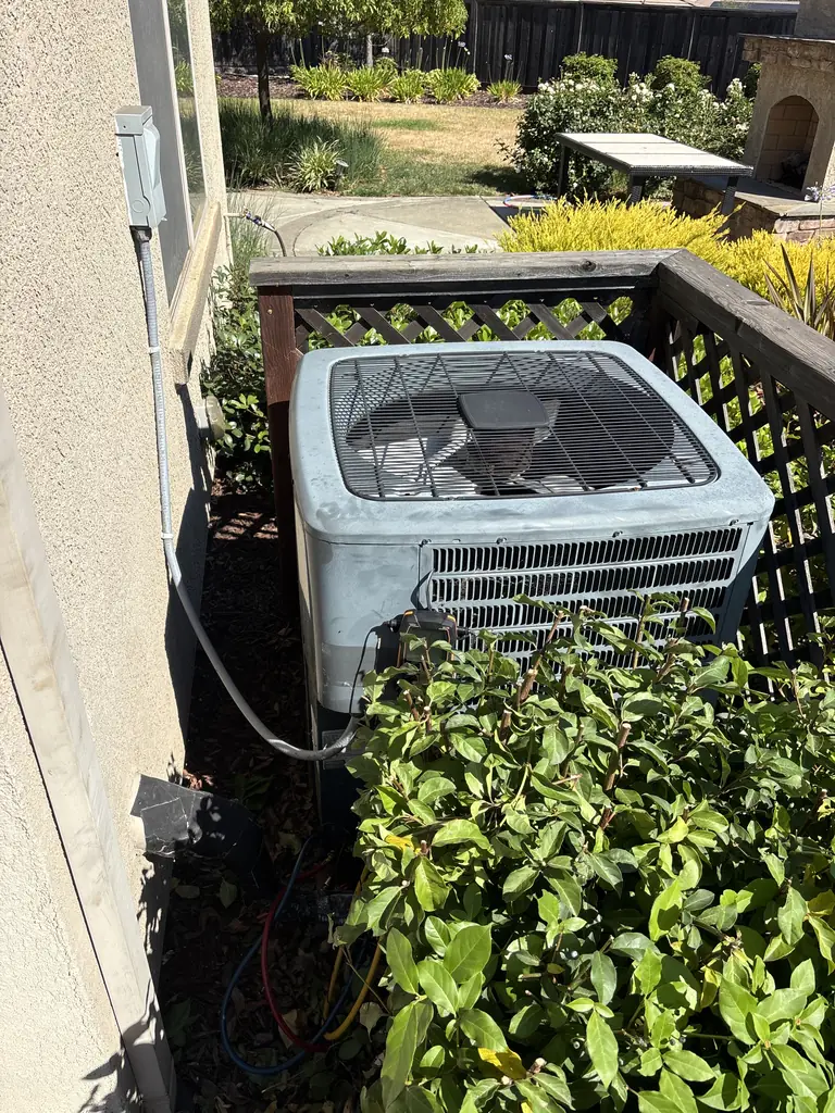 Before: the Goodman R-410A air conditioning condenser beside a Fair Oaks house in June 2026, set against a wood lattice screen with shrubs growing up to the coil and a refrigerant gauge hose connected